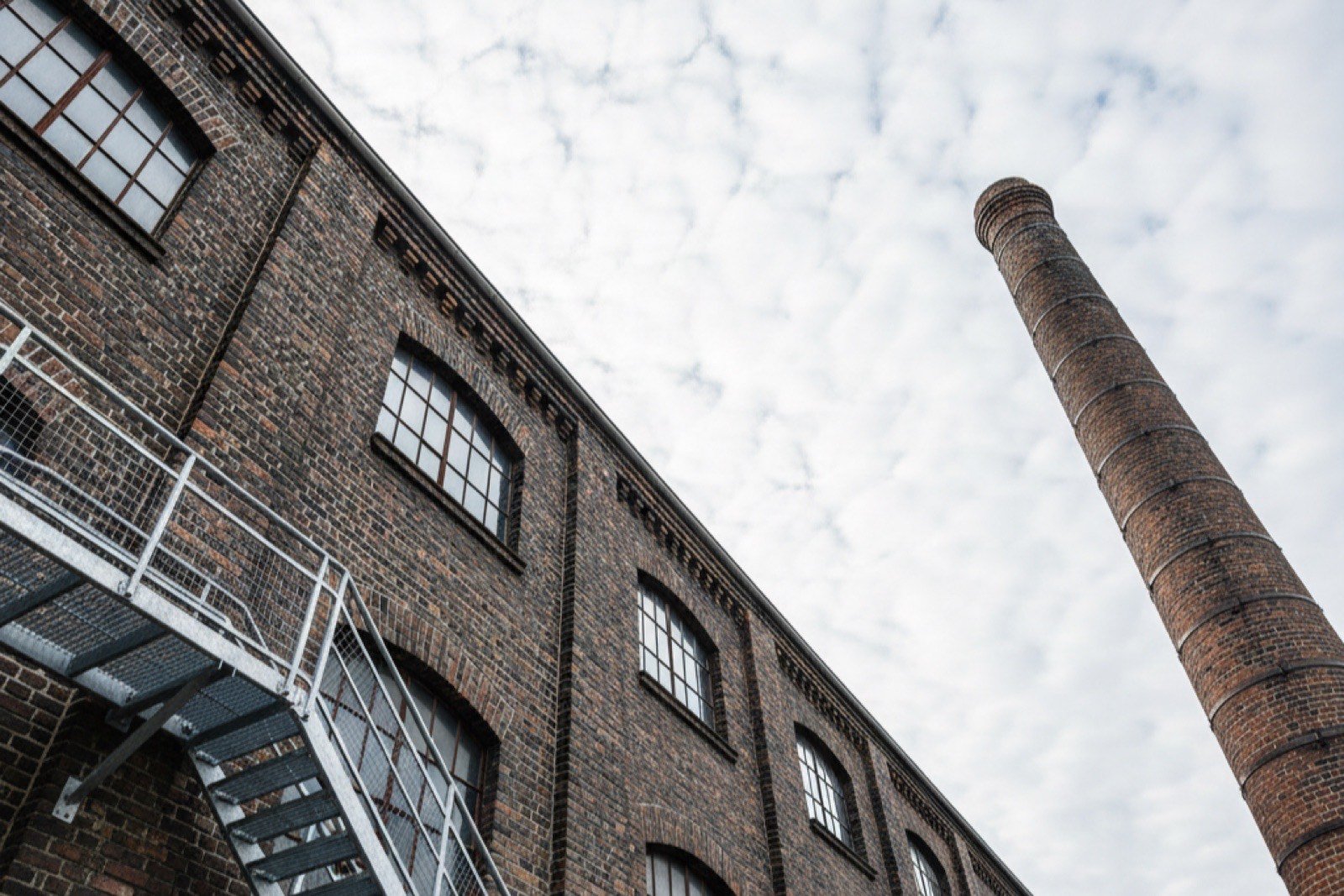 Ältere Backstein-Industriehalle mit hohem Ziegelschornstein und außenliegender Stahltreppe von unten fotografiert
